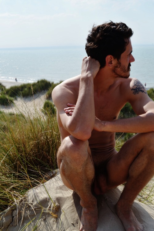 Un homme nu tatoué sur une plage, assis dans les dunes